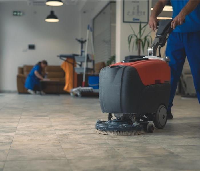 Cleaner cleans hard floor with scrubber machine while other cleaner cleans in the background